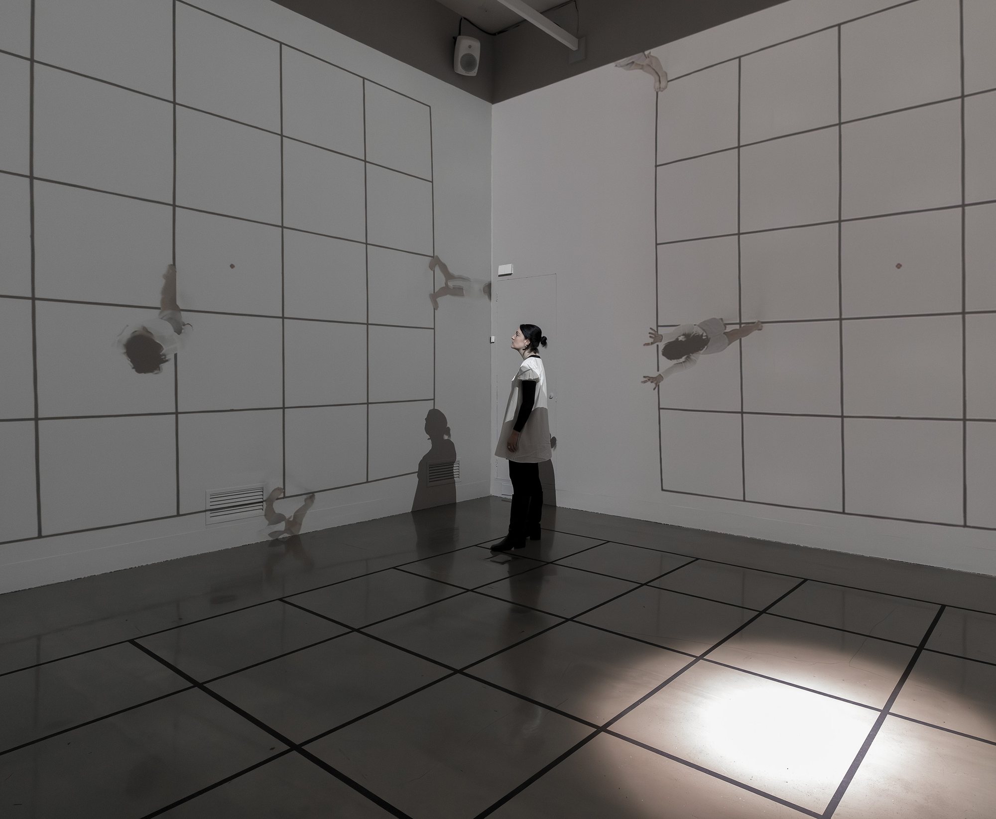 A visitor looks up at dancers floating across two gridded walls, her shadow among them